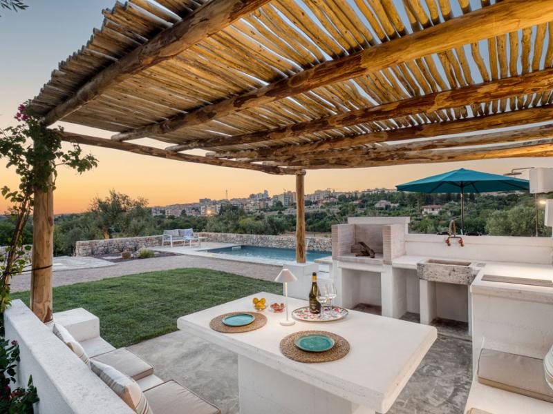 Terrazza con pergola, tavolo da pranzo, barbecue e piscina al tramonto.