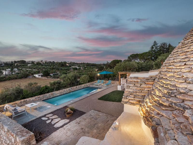 Piscina con vista panoramica su campagna, al tramonto, accanto a una struttura in pietra.